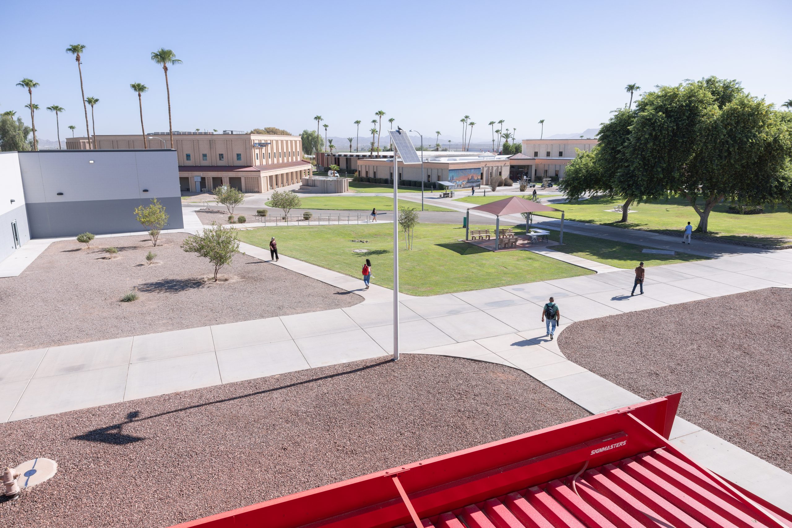 An aerial view of a modern campus with paved walkways, green lawns, palm trees, and buildings under a clear blue sky.